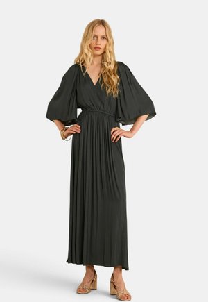 Donna con lunghi capelli biondi che indossa un vestito maxi nero plissettato con maniche larghe e sandali beige con tacco a blocco, in piedi con le mani sui fianchi.