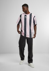 Striped t-shirt featuring light pink and navy blue colors, with a loose fit, short sleeves, and a logo on the front. Paired with dark pants.