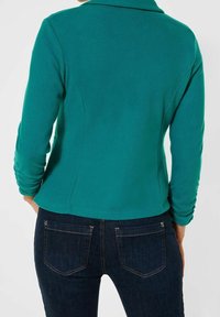 Grön långärmad tröja med krage, figurnära design, ribbad textur och sömligt bakstycke, kombinerad med mörkblå jeans med bakfickor.