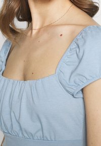Detailní záběr na ženský krk a horní část hrudníku, která nosí světlomodrý top bez ramínek a jemný zlatý náhrdelník.