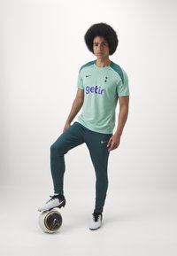 Mint vihreä Nike-urheiluasu purppuralla "getir"-logolla, vihreät korostusolkaimet, yhdistetty tummanvihreisiin housuihin, mustat ja valkoiset nappulakengät, jalka pallolla.