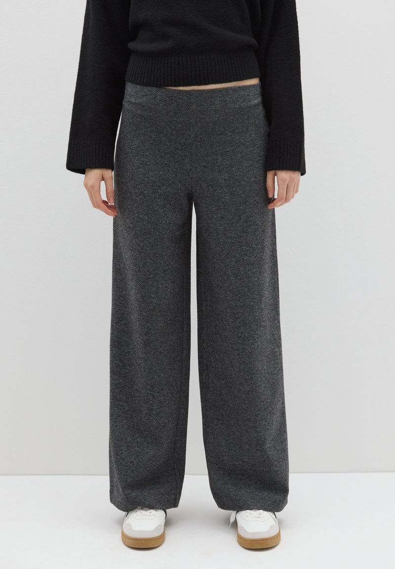 Pantaloni a gamba larga in tessuto grigio scuro con finitura strutturata. Presentano una vita alta e una cintura liscia. Abbinati a sneaker bianche.