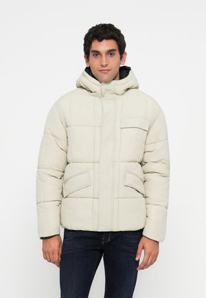 Mand iført en lys beige vatteret hættejakke med klaplommer, stående imod en simpel hvid baggrund.