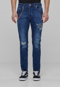Plave traperice s istaknutim isprekidanim detaljima, Slim fit kroj i vidljivim šavovima. Imaju metalni lanac kao detalj i bočne džepove. Uparene su s crnim tenisicama.