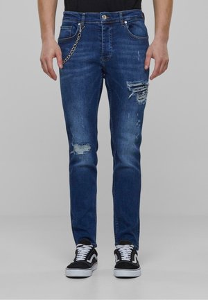 Plave traperice s istaknutim isprekidanim detaljima, Slim fit kroj i vidljivim šavovima. Imaju metalni lanac kao detalj i bočne džepove. Uparene su s crnim tenisicama.