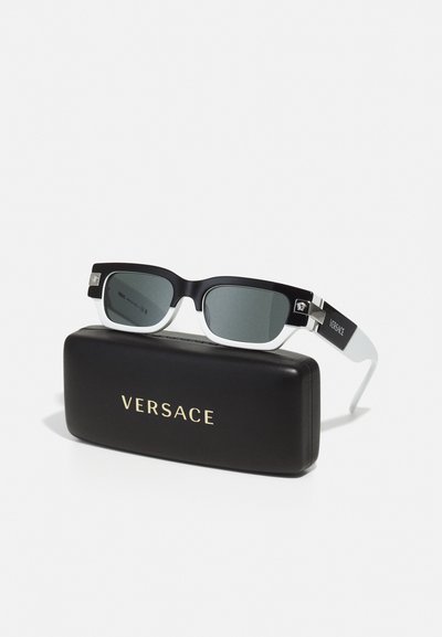 Ochelari de soare rectangulari negri cu lentile gri, având laterale albe și accente aurii, expuși într-o etui Versace negru cu litere aurii.