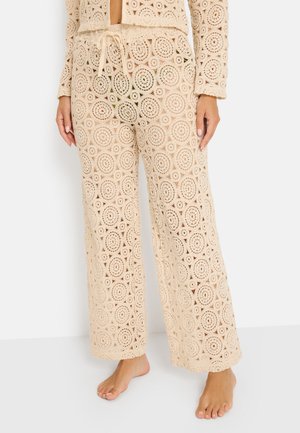 Beige crocheted pants with circular patterns and a drawstring waist, worn barefoot with matching crocheted top visible at waist.