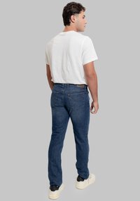 Jeans in denim blu scuro con taglio dritto, cuciture dettagliate e tasche posteriori. Abbinati a una t-shirt bianca semplice e sneakers nere.