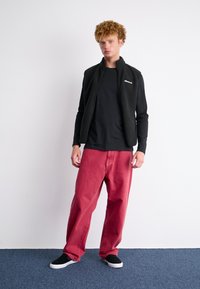 Melnas vilnas veste virs melna garām piedurknēm krekliem, apvienota ar brīvām sarkanām biksēm un melniem kurpēm bez pēdas, izkārtota uz vienkārša fona.