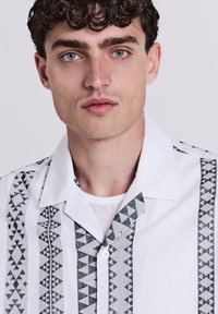 Giovane uomo con capelli ricci scuri e occhi blu che indossa una camicia bianca con motivi geometrici neri su uno sfondo semplice.