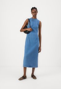 Vestido maxi de mezclilla en azul claro con una silueta ajustada, cuello redondo y detalles de costura sutiles. Llevado con zapatos planos de estampado de leopardo y un bolso negro.