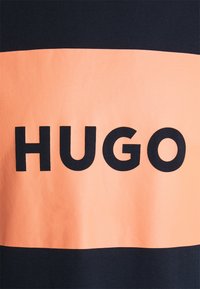 Černé a broskvové bavlněné tričko s výrazným textem "HUGO". Design má horizontální broskvový pruh na černém pozadí. Hladká textura.