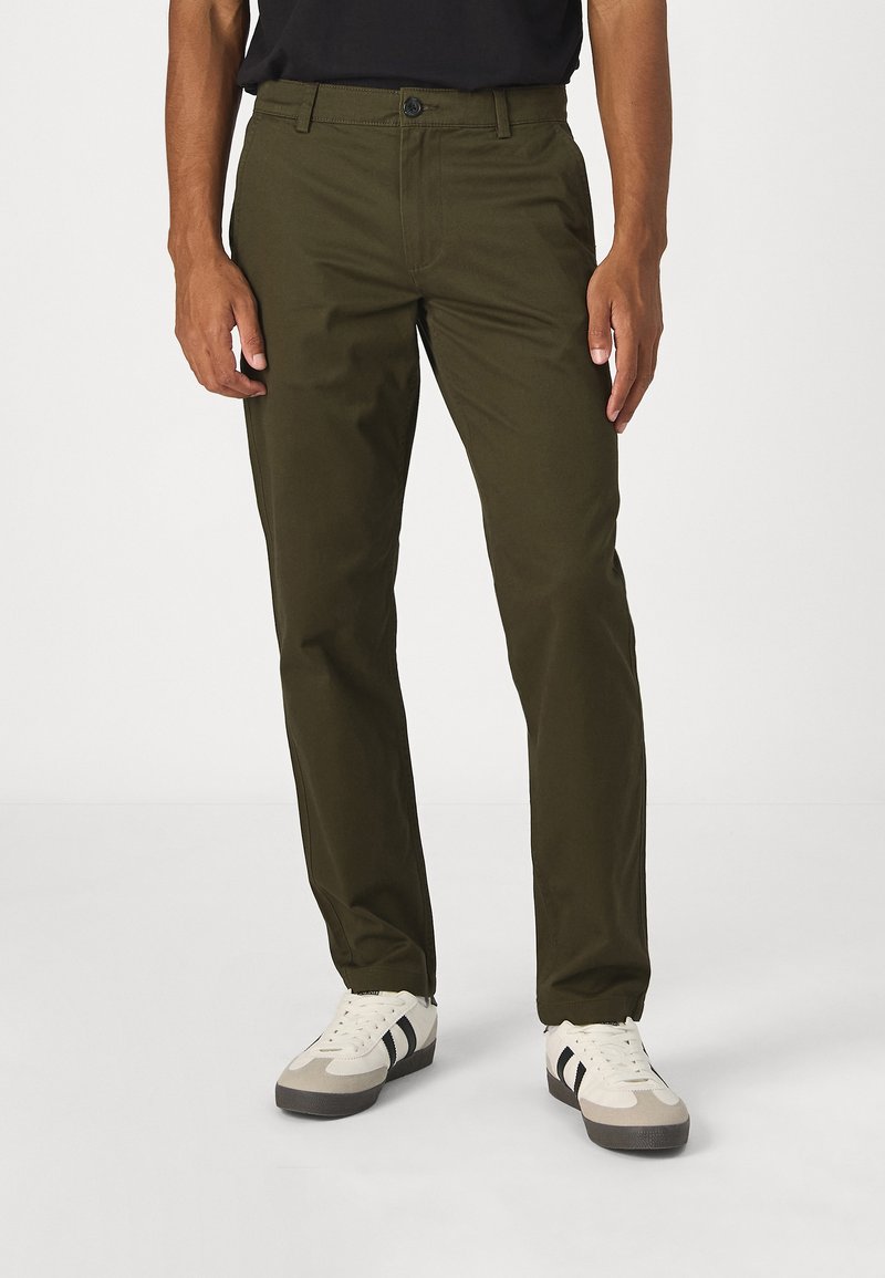 Chinos verde-oliva feitos de um tecido durável. Apresentam um design de perna reta com um clássico fecho de botão e bolsos laterais, usados com ténis brancos.