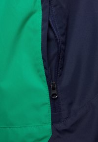 Kurtka w kolorowe bloki z zielonym i granatowym materiałem o gładkiej fakturze, z kieszenią na zamek na granatowym elemencie.