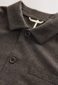 Brown textured polo shirt with a collar, two dark buttons, and a small pocket. Features a woven fabric with a subtle pattern.