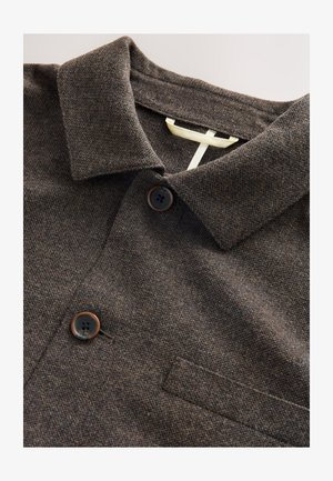 Brown textured polo shirt with a collar, two dark buttons, and a small pocket. Features a woven fabric with a subtle pattern.