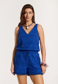 Plavi kombinezon s uzorkom, V-izrezom, elastičnom strukom i resama na rubu. Dodaci uključuju narukvicu i ogrlicu od višeobojenih perli.