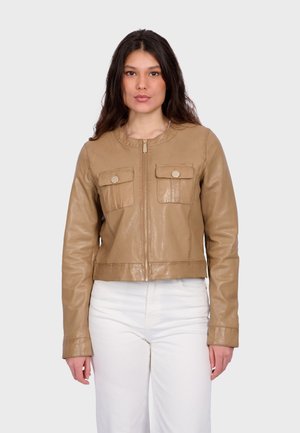 Femme aux longs cheveux noirs portant une veste en cuir marron clair avec des poches avant et un pantalon blanc, debout devant un fond clair uni.