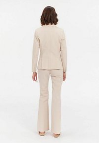 Costume beige ajusté avec une veste cintrée et un pantalon évasé. Tissu texturé, fermeture à un bouton et détails de couture subtils.