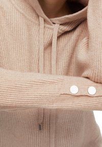 Pull beige en tricot avec capuche, cordons de serrage et deux boutons blancs sur la manche repliée.