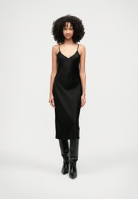 Sieviete ar cirtainiem matiem, tērpta melnā zīda kleitā un melnās augstajās zābakos, stāv uz vienkārša baltā fona.