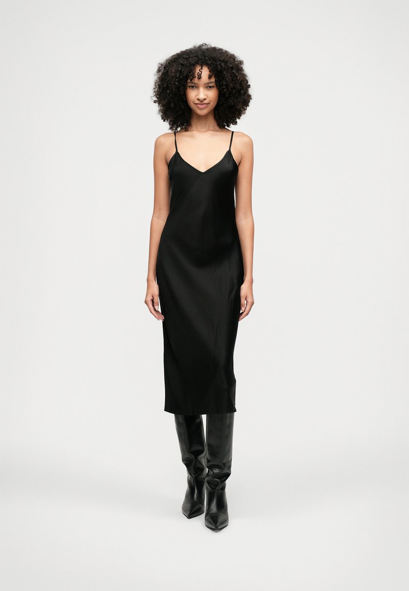 Sieviete ar cirtainiem matiem, tērpta melnā zīda kleitā un melnās augstajās zābakos, stāv uz vienkārša baltā fona.