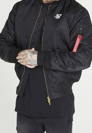 Czarna kurtka bomber z złotym zamkiem błyskawicznym, białe logo na lewej piersi, czerwona metka "SikSilk" na kieszeni rękawa, noszona na czarnej koszuli, splecione dłonie, tatuaże.