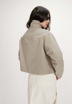 Femme aux longs cheveux foncés portant des lunettes de soleil noires et une veste beige courte sur une jupe blanche, vue de profil sur un fond uni.