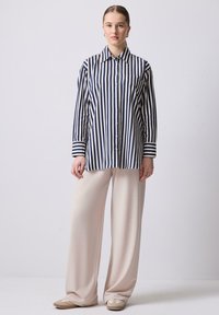 Camicia a righe blu navy e crema con colletto, maniche lunghe e tasca sul petto, abbinata a pantaloni a gamba larga beige chiaro.