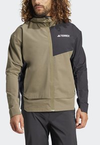 Man in een twee-kleurige groen en zwarte Terrex hoodie-jack en zwarte broek, staande tegen een lichtgrijze achtergrond.