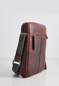 Hnedá kožená crossbody taška s prednou zipsovou kapsou a nastaviteľným čierno-bielym tkaným popruhom na jednoduchom pozadí.