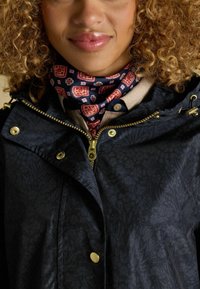 Giacca blu navy con motivo floreale, chiusura frontale con zip, dettagli in oro e cappuccio, abbinata a una sciarpa con motivo nei toni del blu e del rosso.