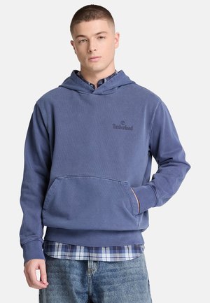 Junger Mann, der einen blauen Timberland-Hoodie über einem blau karierten Hemd mit Jeans trägt und mit einer Hand in der Kapuzen-Tasche steht.