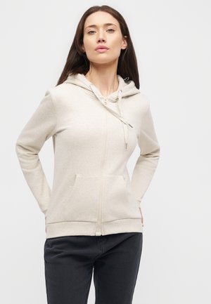 Une femme porte un sweat à capuche crème zippé, avec des poches sur le devant, et un pantalon foncé sur un fond clair et uni.