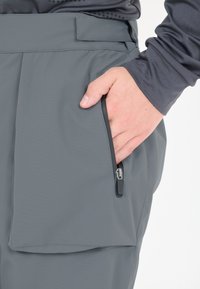 Grijze stoffen shorts hebben een zijzak met ritssluiting en een gladde textuur. De tailleband is voorzien van een klittenbandsluiting voor een verstelbare pasvorm.