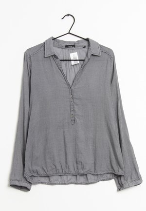 Blouse grise à manches longues avec petits pois noirs, un col et des boutons, suspendue sur un cintre noir sur fond blanc.