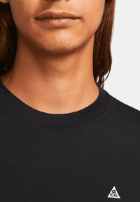 Svart bomull t-shirt med rund hals, ribbad krage och en liten vit triangel-logotyp på bröstet. Slät textur.