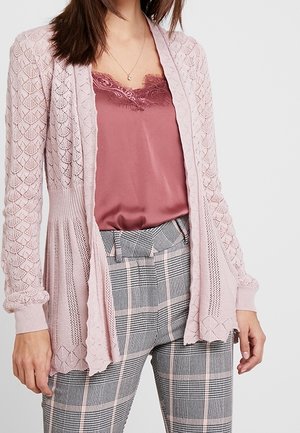 Vrouw draagt een stoffig roze hemdje met kanten rand, een lichtroze gebreid vest en een grijze geruite broek met hoge taille en roze strepen.