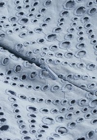 Tissu bleu clair avec des motifs en œillets brodés, présentant des ouvertures circulaires et un détail de fermeture éclair. Texture lisse, design délicat.