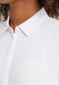 Close-up van een persoon in een witte blouse met opgerolde kraag, met zicht op de nek en een deel van krullend haar.