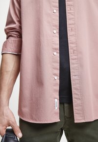Camicia button-up rosa chiaro con bottoni bianchi, maniche corte e una leggera texture, abbinata a una camicia nera e pantaloni verde oliva.