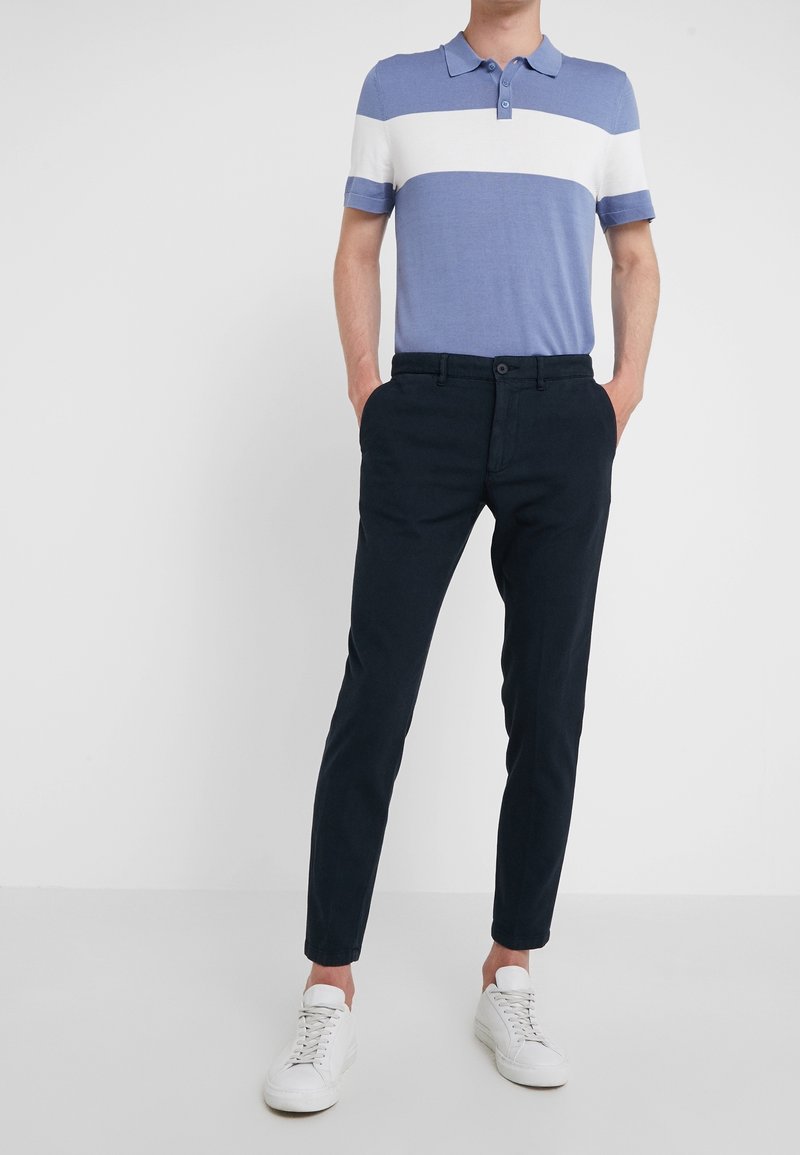 Námornické slim-fit kalhoty s lehkým polo tričkem modré a bílé pruhy. Model má na sobě bílé tenisky a stojí na jednobarevném pozadí.