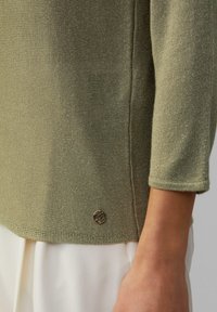 Jasnozielony sweter z dzianiny z rękawami o długości 3/4, z małym, złotym logo przy dolnym brzegu. Miękka tekstura i delikatne prążkowanie.