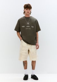 T-shirt oversize verde oliva con testo grafico, abbinata a pantaloni corti tie-dye color crema. Scarpe nere, calzini bianchi. Il modello è in piedi contro uno sfondo semplice.