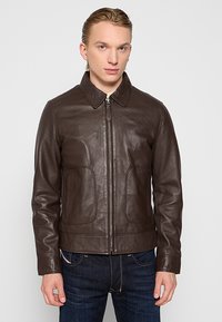 Giacca di pelle marrone con colletto, zip frontale e due tasche laterali. Texture liscia e design aderente, abbinata a jeans in denim scuro.