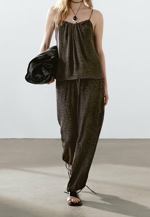 Woman wearing black patterned sleeveless top and loose pants, holding a black clutch bag, with strappy black sandals and layered necklaces.