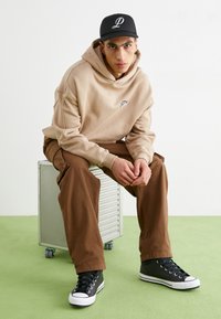 Beige hooded sweatshirt with logo, brown cargo pants, and black high-top sneakers with white accents. Seated on a metallic rolling cart.