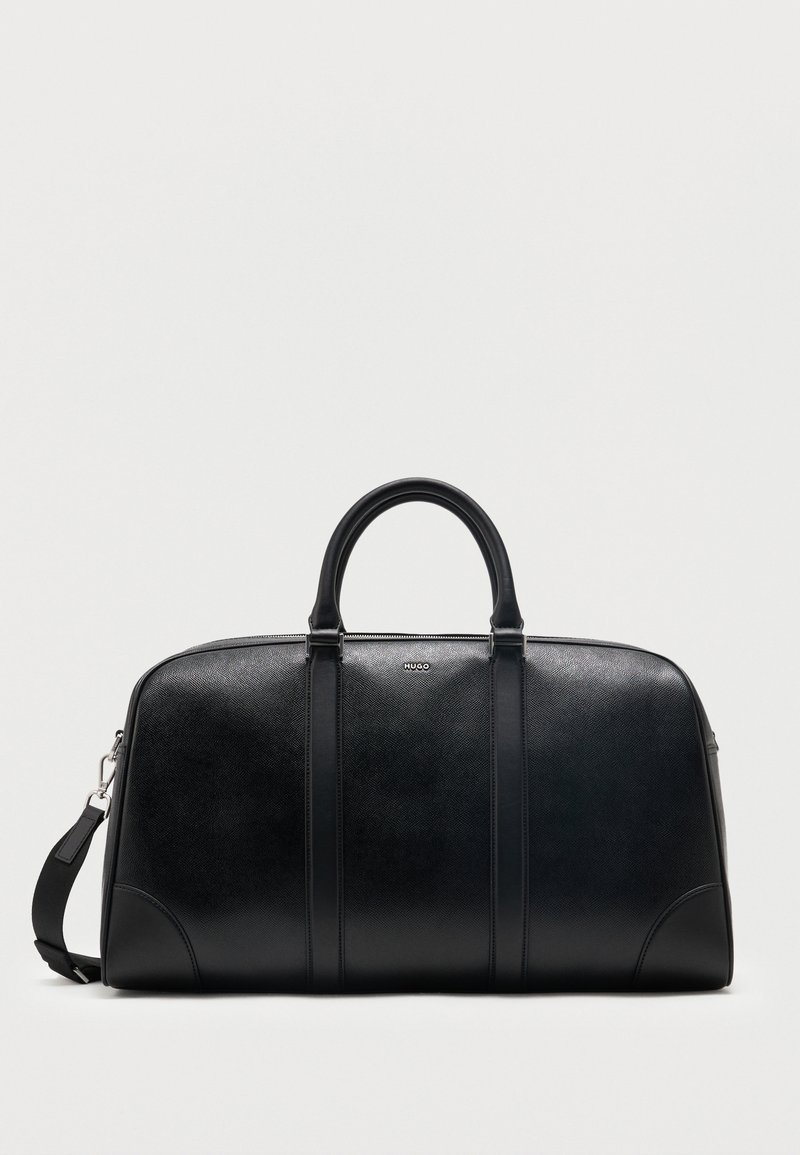 Sac de voyage en cuir noir avec deux poignées supérieures, bandoulière amovible et petit logo "Hugo" centré près de la fermeture éclair supérieure.