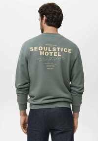 Green sweatshirt with a crew neckline and ribbed cuffs. Back features large cream text "SEOULSTICE HOTEL" and smaller hotel details below.