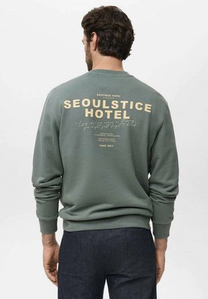 Sweatshirt vert avec un col rond et des poignets côtelés. Le dos présente un grand texte crème "SEOULSTICE HOTEL" et des détails plus petits de l'hôtel en dessous.